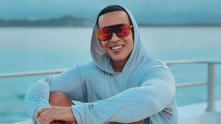 "Daddy Yankee sonriendo en un yate con gafas de sol rojas y sudadera con capucha gris."